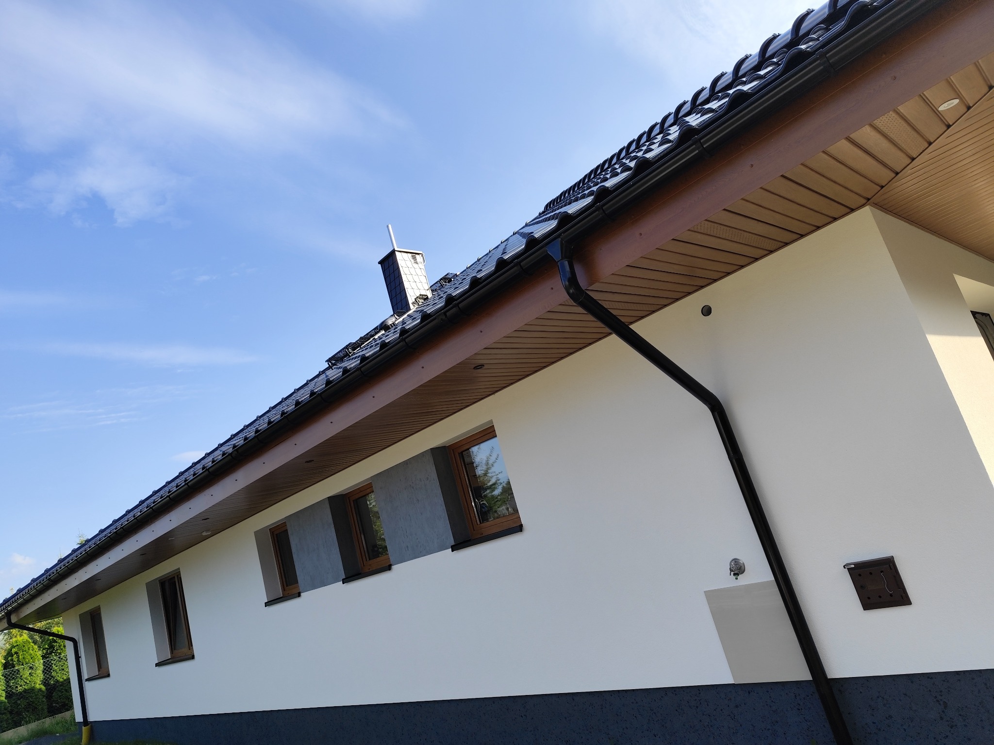 Nowoczesny dom jednorodzinny z białą elewacją, fragmentem tynku strukturalnego w odcieniach szarości wokół okien, ciemną podmurówką, brązowym podbitką dachową i czarną rynną, sfotografowany...