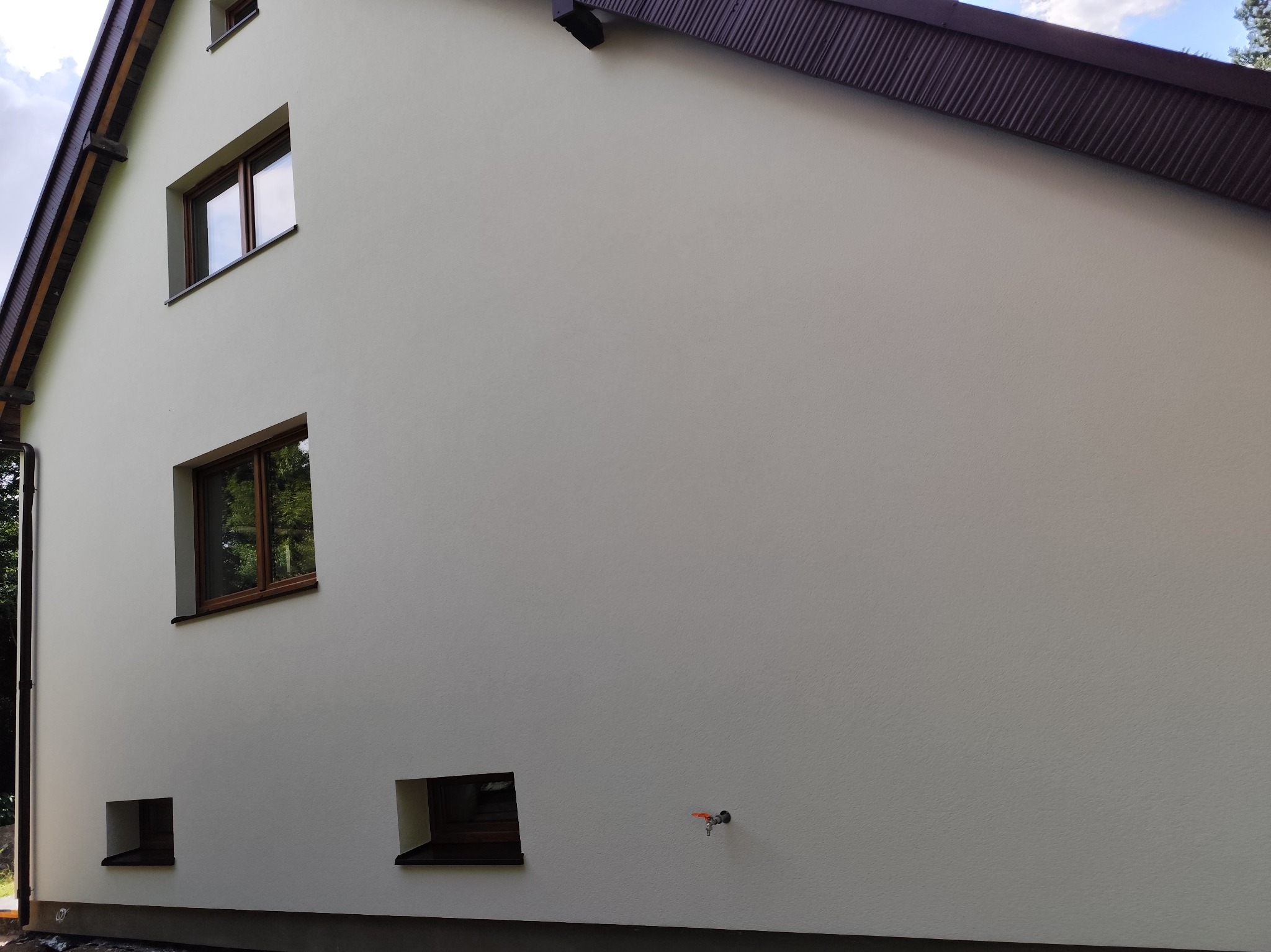 Jasna elewacja domu jednorodzinnego z brązowymi ramami okiennymi i ciemnobrązowym dachem, widoczne trzy okna na różnych wysokościach i kran na ścianie.