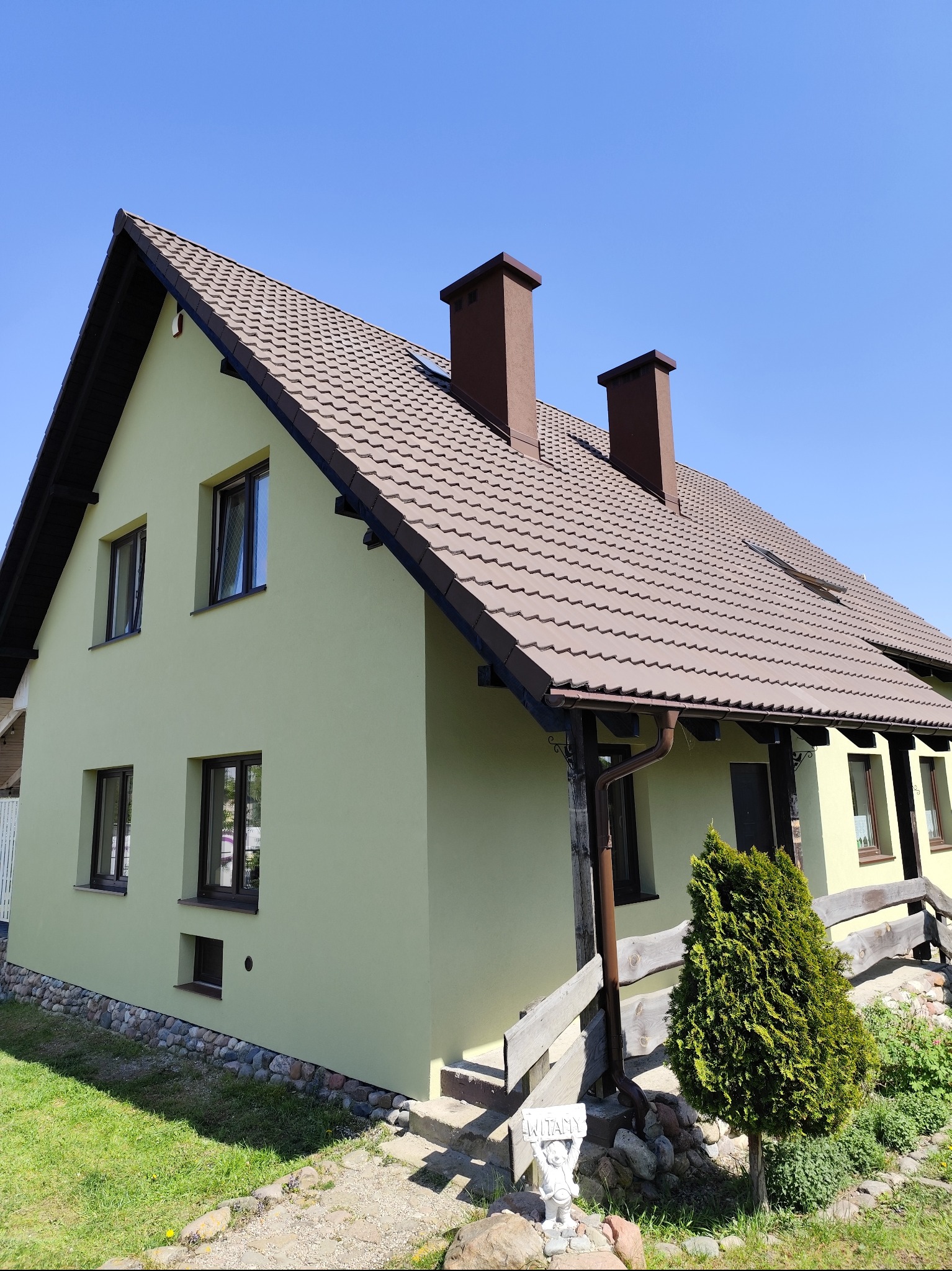 Dom jednorodzinny z jasną elewacją o strukturze baranka, brązową dachówką, ciemnymi oknami i kominami na tle błękitnego nieba, z widocznym fragmentem ogrodu z kamienną ścieżką i dekoracyjną figurką