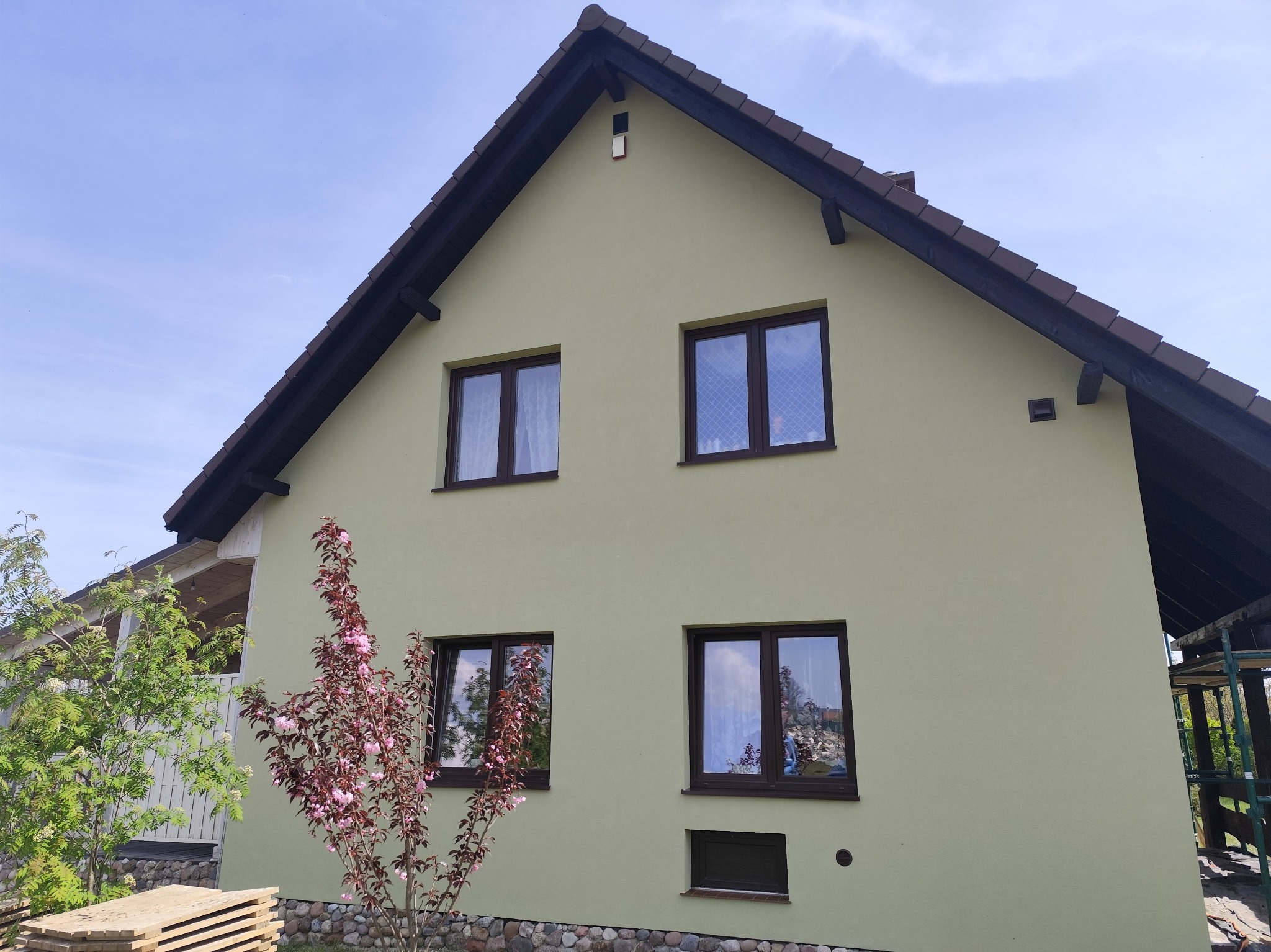 Dom jednorodzinny z jasną elewacją o strukturze baranka, ciemną stolarką okienną i brązowym dachem, widoczny fragment dekoracyjnego drzewa na pierwszym planie.