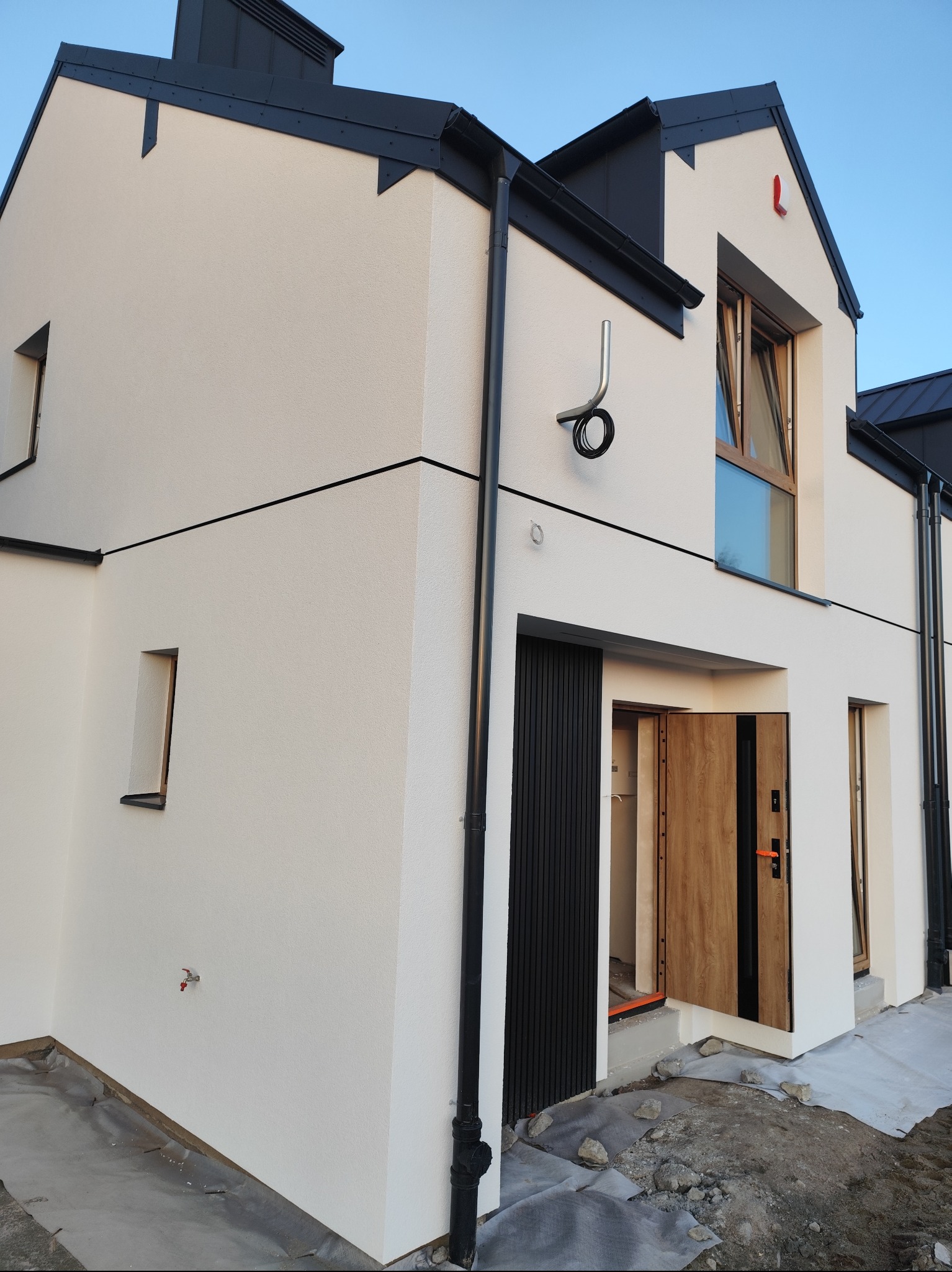 Nowoczesny dom jednorodzinny z jasną elewacją, czarnymi akcentami dachu i stolarki okiennej, widoczne elementy wykończeniowe i teren wokół budynku w trakcie prac.