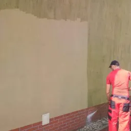 Pracownik w czerwonym kombinezonie czyści elewację budynku z ceglaną podmurówką za pomocą myjki ciśnieniowej, widoczna różnica w czystości ściany.