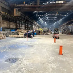 Grupa osób w kaskach podczas przerwy w hali produkcyjnej z wózkiem widłowym Linde w tle, widoczne palety i elementy konstrukcyjne hali.