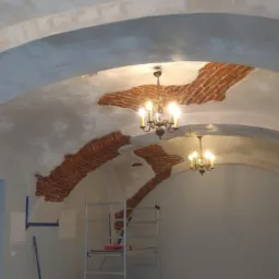 Wnętrze z łukowym sufitem w trakcie prac wykończeniowych, widoczne fragmenty odsłoniętej cegły, częściowo pokryte gładzią, metalowa drabina i wiszące żyrandole.