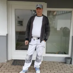 Mężczyzna w ubraniu roboczym, z papierosem w dłoni, stoi przed budynkiem z pomalowaną elewacją i oknami.