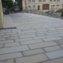 Świeżo ułożona, jasnoszara nawierzchnia z prostokątnych elementów, widoczny budynek w tle i sprzęt budowlany za ogrodzeniem.