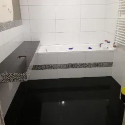 Częściowo wykończona łazienka z czarną podłogą i blatem, białe płytki ścienne z dekoracyjnym pasem, wanna zabezpieczona folią, grzejnik drabinkowy.