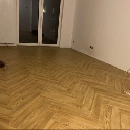 Droz-Bud - Jasna podłoga w jodełkę z drewnopodobnych paneli w pustym pokoju z białymi ścianami, dużymi drzwiami balkonowymi i białym grzejnikiem. Widoczne narzędzia przy ścianie.