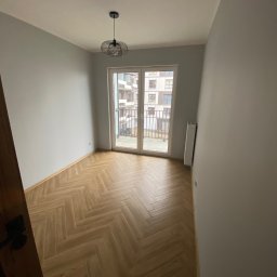 Droz-Bud - Jasny pokój z podłogą w jodełkę, świeżo pomalowanymi szarymi ścianami i nowoczesną lampą wiszącą. Duże drzwi balkonowe z widokiem na osiedle.