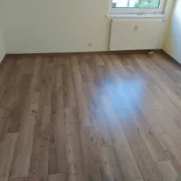 Wykończona podłoga z paneli w odcieniach brązu, widok na grzejnik i gniazdko elektryczne w odnowionym pokoju we Wrocławiu.