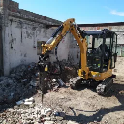 Żółta mini koparka CAT z młotem hydraulicznym rozbija gruz na tle zniszczonego budynku z cegły i błękitnego nieba.