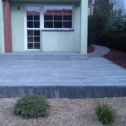 Nowoczesny taras z szarej kostki brukowej, obrzeża z ciemniejszych kostek, żwirowe podłoże i nasadzenia z niskich krzewów, widok na dom z oknem i schodami.