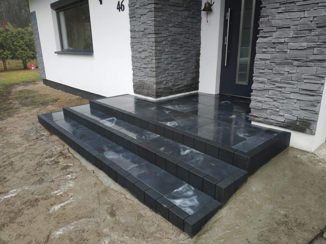 Nowoczesne schody wejściowe z ciemnej kostki granitowej, wykończone obrzeżem z kostki brukowej, prowadzące do drzwi wejściowych domu z elewacją z kamienia dekoracyjnego.