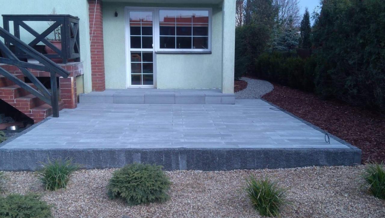 Nowoczesny taras z szarej kostki brukowej, obrzeża z ciemniejszych kostek, żwirowe podłoże i nasadzenia z niskich krzewów, widok na dom z oknem i schodami.
