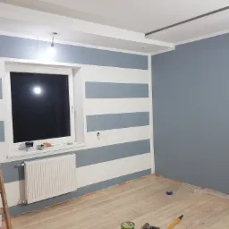 Pokój w trakcie malowania: ściana w poziome pasy (szare i białe), druga ściana szara, podłoga zabezpieczona folią, widoczna drabina malarska, puszka z farbą na parapecie.