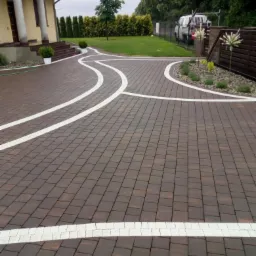 Nowoczesna posesja z brązową kostką brukową oddzieloną białymi pasami, prowadząca do domu z kolumnami i zadbanym trawnikiem z krzewami.