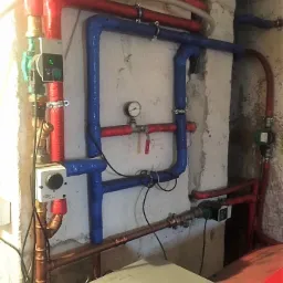 Złożona instalacja hydrauliczna z rurami w kolorze czerwonym i niebieskim, izolowanymi taśmą, widoczny manometr i sterowniki, zamontowana na surowej, betonowej ścianie.