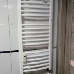 Biały, drabinkowy grzejnik łazienkowy zamontowany na ścianie z białych płytek, z widocznym termostatem i zaworami, obok drzwi z drewnopodobną okleiną.