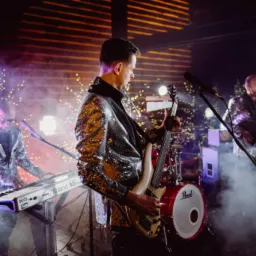 Trzech muzyków w błyszczących marynarkach gra na scenie oświetlonej dekoracyjnymi lampkami i dymem: basista na pierwszym planie, klawiszowiec po lewej i gitarzysta w tle.