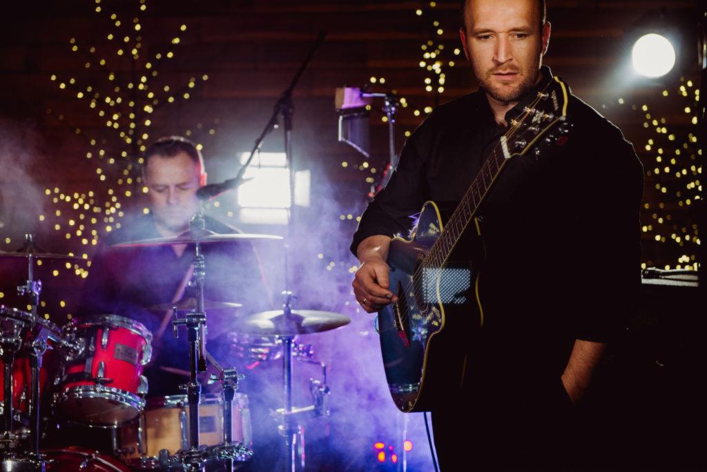 Gitarzysta w czarnej koszuli, w tle perkusista i dekoracje świetlne, scena oświetlona fioletowym światłem.