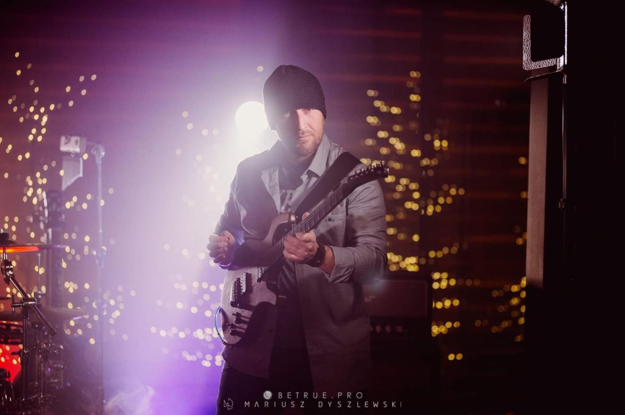 Gitarzysta w czapce, z elektryczną gitarą na scenie, z efektem świetlnym i perkusją w tle.