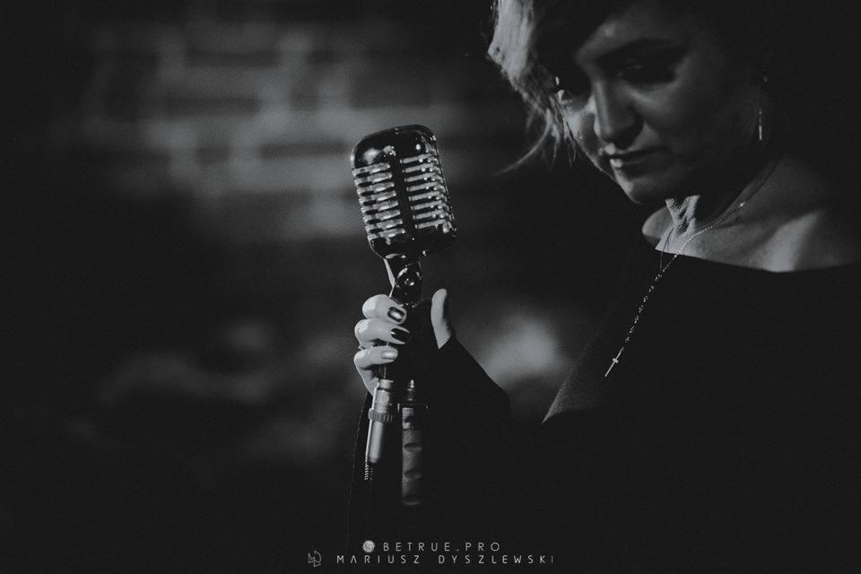 Czarno-białe zdjęcie piosenkarki trzymającej mikrofon w stylu retro, z delikatnym uśmiechem i widocznym łańcuszkiem z krzyżykiem na szyi.