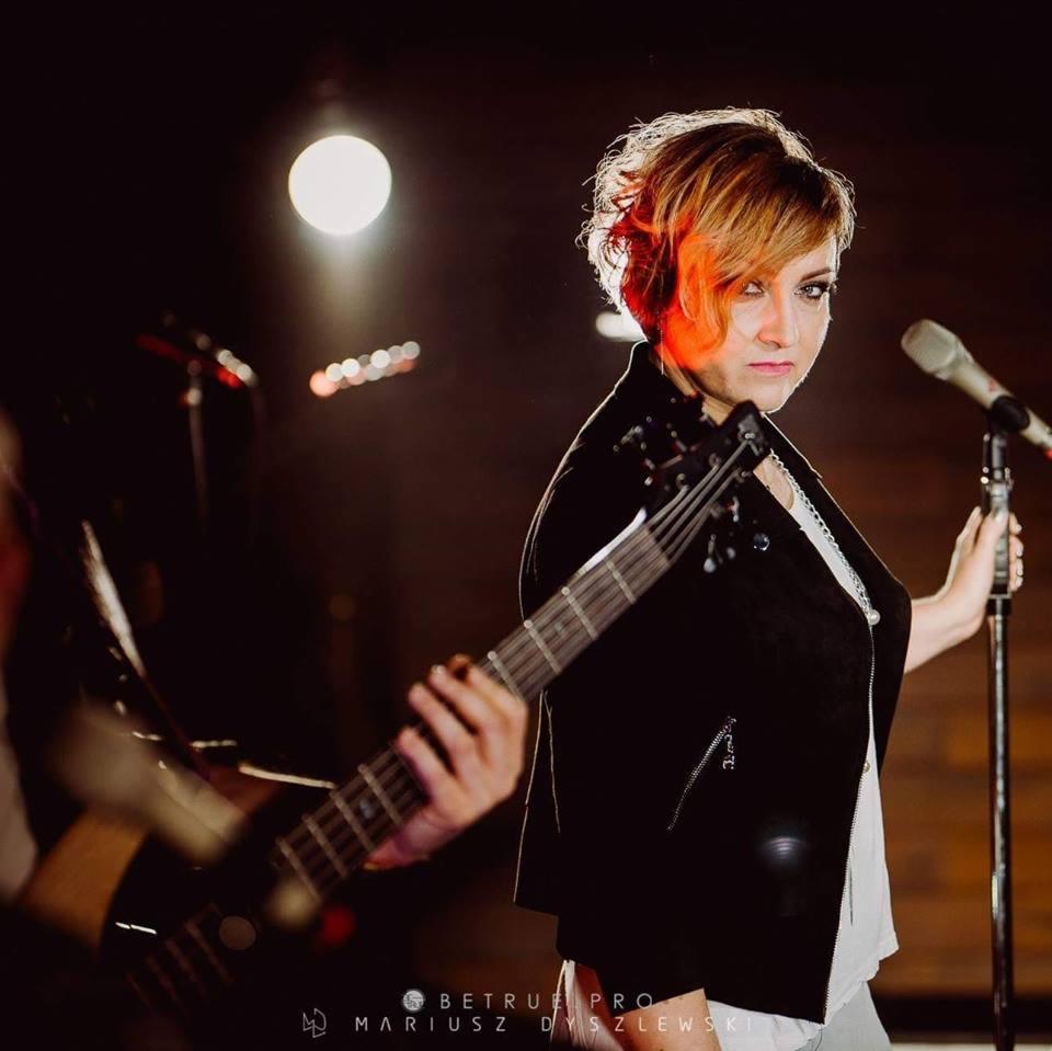 Kobieta z krótkimi włosami w czarnej kurtce, trzymająca gitarę basową i mikrofon na scenie z ciemnym tłem i reflektorami.