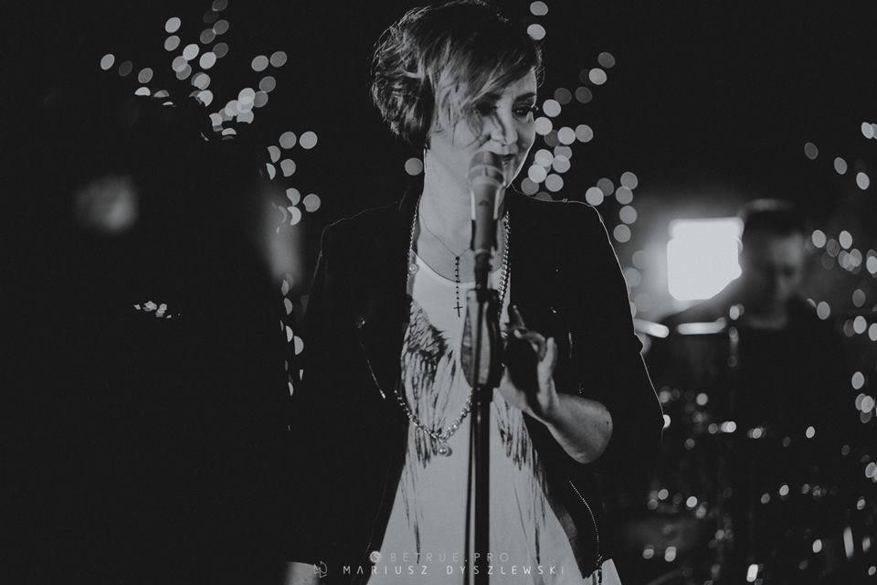Czarno-białe zdjęcie wokalistki z krótkimi włosami, śpiewającej do mikrofonu na scenie z rozmytymi światłami w tle, w tle widoczny perkusista.