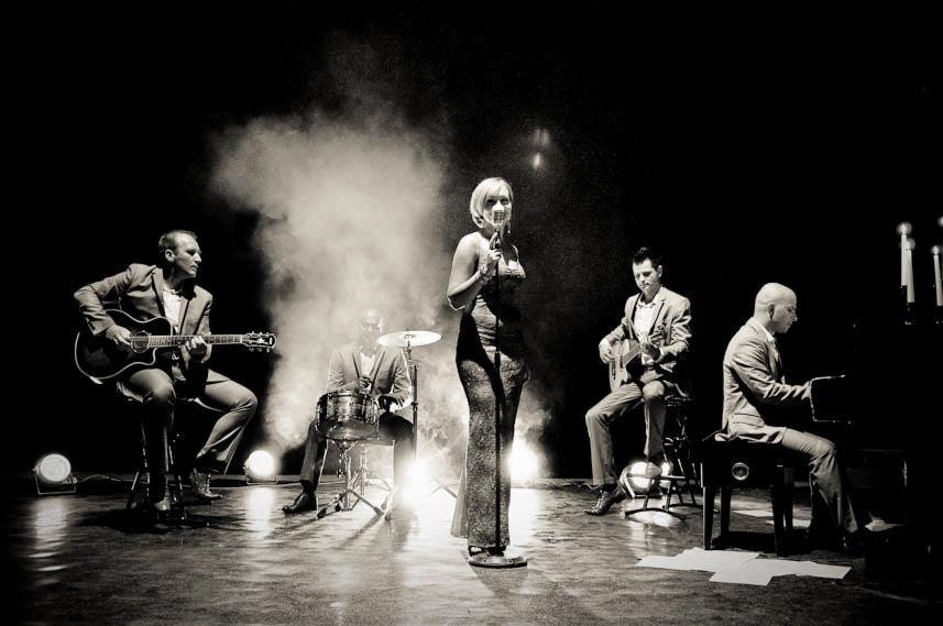 Czarno-białe zdjęcie zespołu muzycznego na scenie, wokalistka w błyszczącej sukni, gitarzyści, perkusista i pianista w garniturach, oświetlenie sceniczne z efektem dymu.