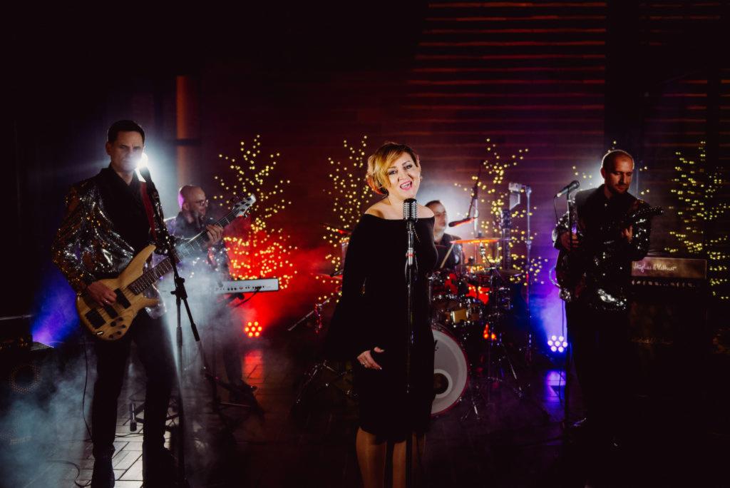 Zespół muzyczny na scenie: wokalistka w czarnej sukience śpiewa do mikrofonu, obok gitarzysta w cekinowej marynarce, w tle perkusja i dekoracje świetlne.