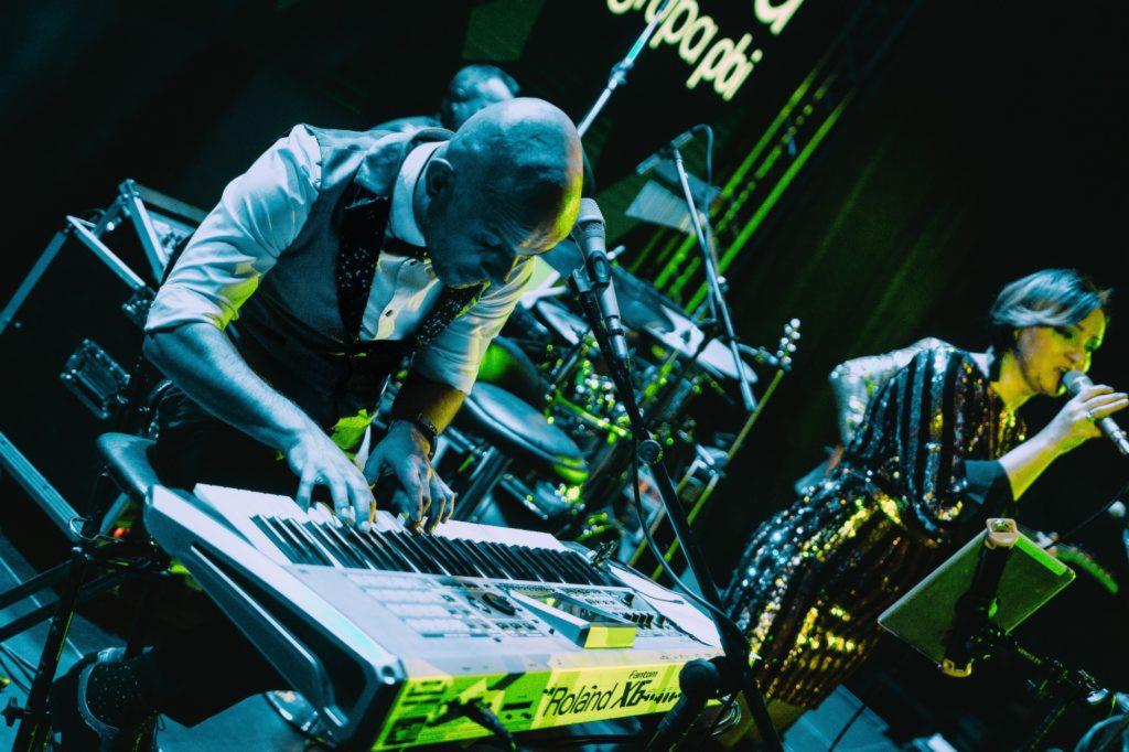 Keyboardzista gra na klawiszach Roland podczas występu na scenie, wokalistka w cekinowej sukience śpiewa do mikrofonu, w tle perkusja i oświetlenie sceniczne.