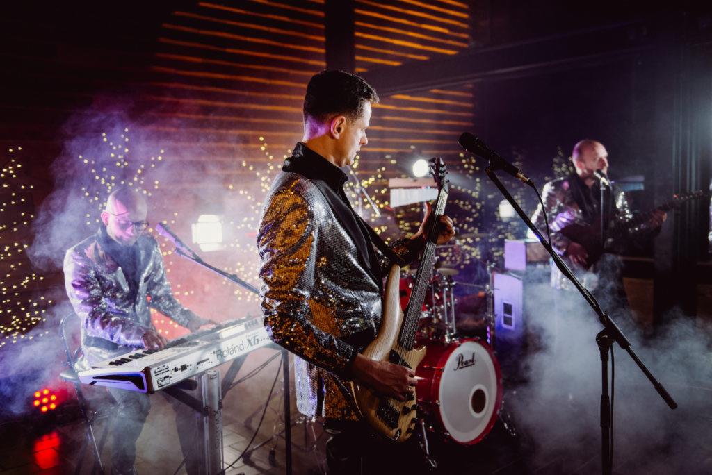 Trzech muzyków w błyszczących marynarkach gra na scenie oświetlonej dekoracyjnymi lampkami i dymem: basista na pierwszym planie, klawiszowiec po lewej i gitarzysta w tle.