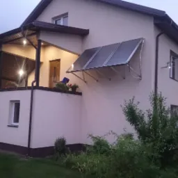 Ocieplona elewacja domu z zamontowanymi panelami słonecznymi nad balkonem, widoczne oświetlenie zewnętrzne i roślinność w ogrodzie.