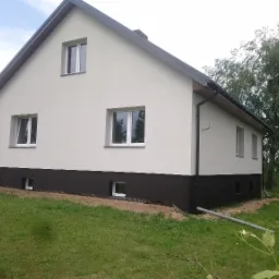 Dom jednorodzinny po wykonaniu elewacji w kolorze białym z ciemnobrązowym cokołem, widoczny fragment ogrodu i błękitny samochód dostawczy w tle.