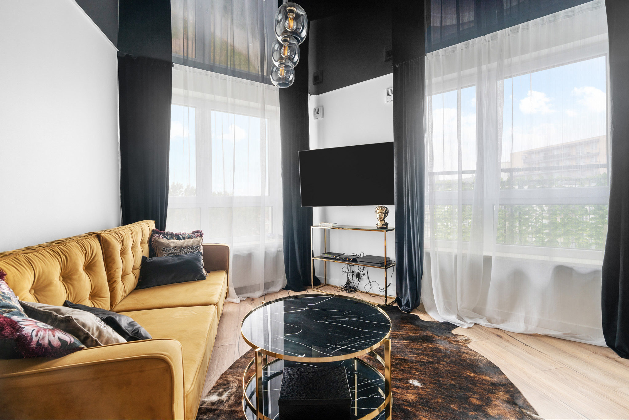 Nowoczesny salon z musztardową sofą, okrągłym stolikiem kawowym z czarnym marmurowym blatem i złotą podstawą, telewizorem na ścianie i dywanem imitującym skórę krowy.