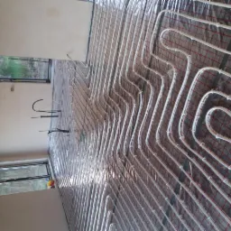 Białe rury ogrzewania podłogowego ułożone na folii z czerwoną siatką, widok na surową podłogę i okna z widokiem na zieleń.