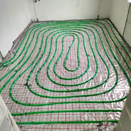 Zielone rury ogrzewania podłogowego ułożone na folii z czerwoną siatką w surowym wnętrzu, widoczne wystające kable elektryczne i fragment ściany z niedokończonym tynkiem.