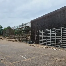 Przygotowanie szalunków zbrojeniowych na placu budowy, widoczne rusztowania i składowane drewno w tle.