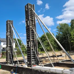 Szare szalunki pionowe wzmocnione metalowymi podporami ukośnymi, przygotowane do wylania betonu na fundamencie budynku w słoneczny dzień. W tle widoczne drzewa i fragment zabudowy mieszkalnej.
