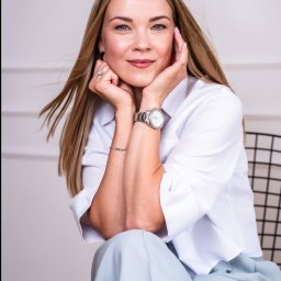 Adwokat Julita Kopeć - Portret uśmiechniętej kobiety w białej koszuli, siedzącej na krześle. Na ręce srebrny zegarek i bransoletka. W tle biała ściana z delikatną sztukaterią.