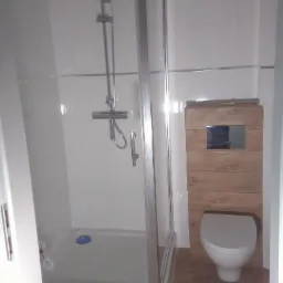 Nowoczesna łazienka z kabiną prysznicową z chromowanymi profilami, toaletą wiszącą z drewnianą obudową i białymi płytkami na ścianach.