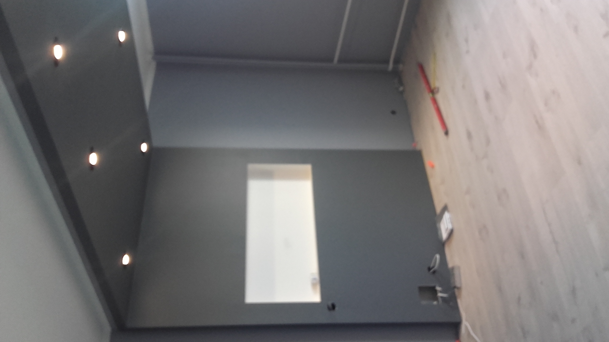 Szara ściana z wnęką, drewnopodobna podłoga, poziomica oparta o ścianę, widoczne punkty świetlne na suficie.