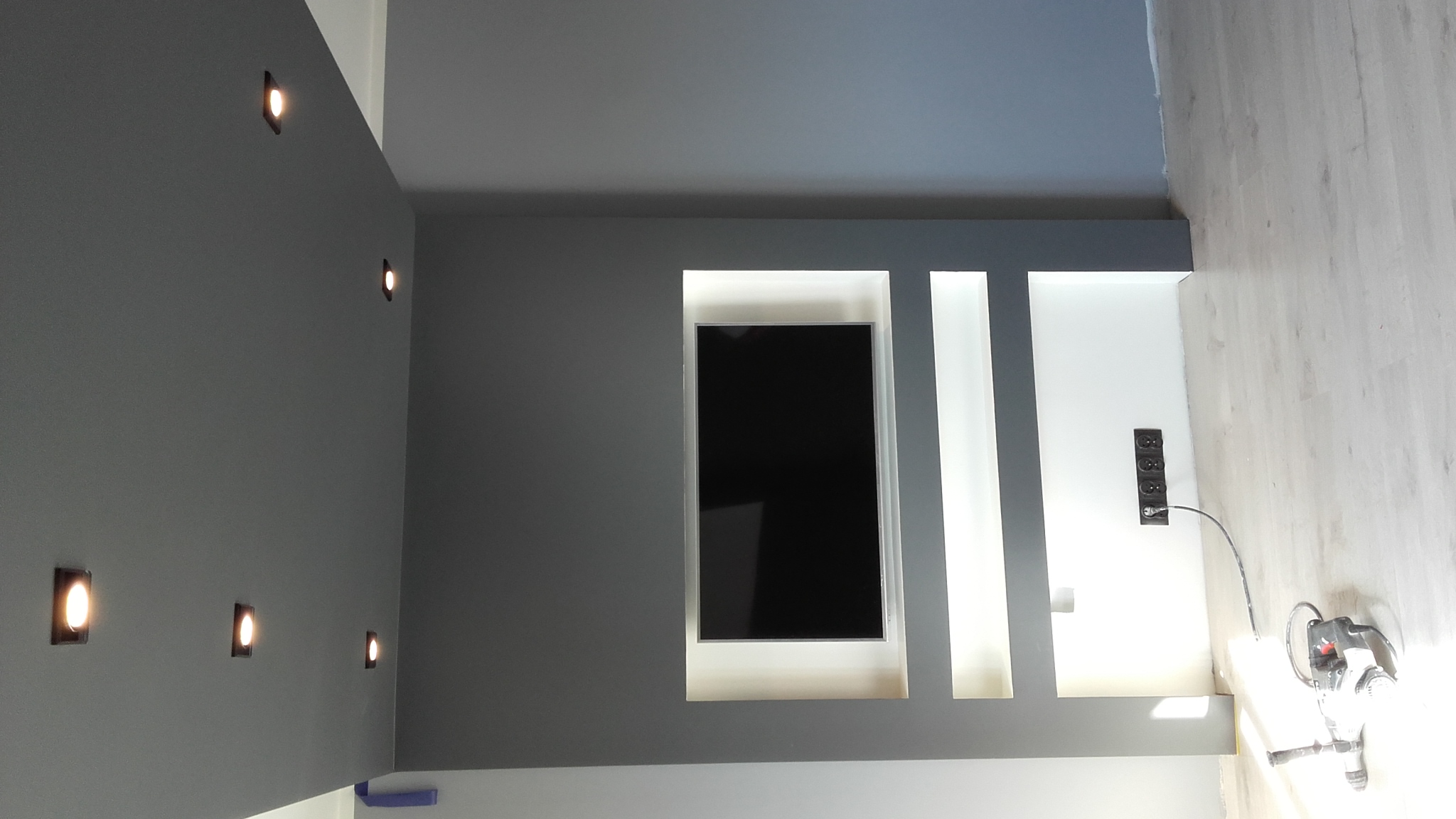 Szara ściana z wnęką na telewizor, punktowe oświetlenie sufitowe i gniazdka elektryczne, widoczna szlifierka do gładzi.