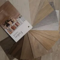 Prezentacja wachlarza próbek paneli podłogowych marki Fusion Wood w różnych odcieniach brązu, beżu i szarości, ułożonych na podłodze z podobnym wzorem drewna.