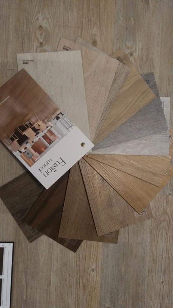 Prezentacja wachlarza próbek paneli podłogowych marki Fusion Wood w różnych odcieniach brązu, beżu i szarości, ułożonych na podłodze z podobnym wzorem drewna.