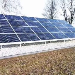 Instalacja fotowoltaiczna na gruncie, panele umieszczone na białym żwirze, widoczne drzewa w tle.