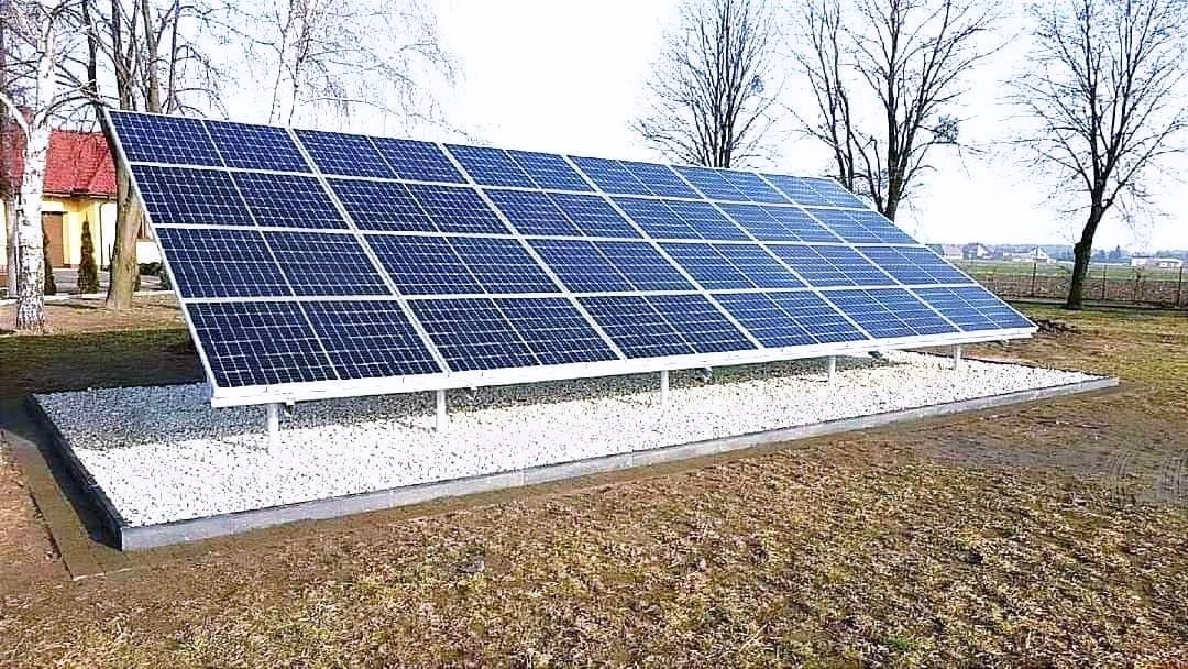 Instalacja fotowoltaiczna na gruncie, panele umieszczone na białym żwirze, widoczne drzewa w tle.