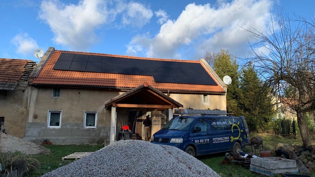 Częściowy montaż paneli na dachu starego domu z czerwonej dachówki, widoczny firmowy bus na trawniku i sterta gruzu, fragment dachu w trakcie remontu, pochmurne niebo.