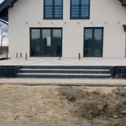 Nowoczesny taras z betonowych płyt i schodami przed domem z dużymi oknami, widoczne przygotowania do dalszych prac ziemnych.