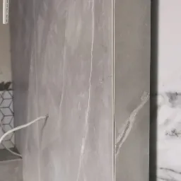 Ściana wyłożona szarymi płytkami imitującymi marmur z widocznymi białymi żyłami, obok fragment ściany z białymi płytkami, instalacja pod baterię wystająca ze ściany.